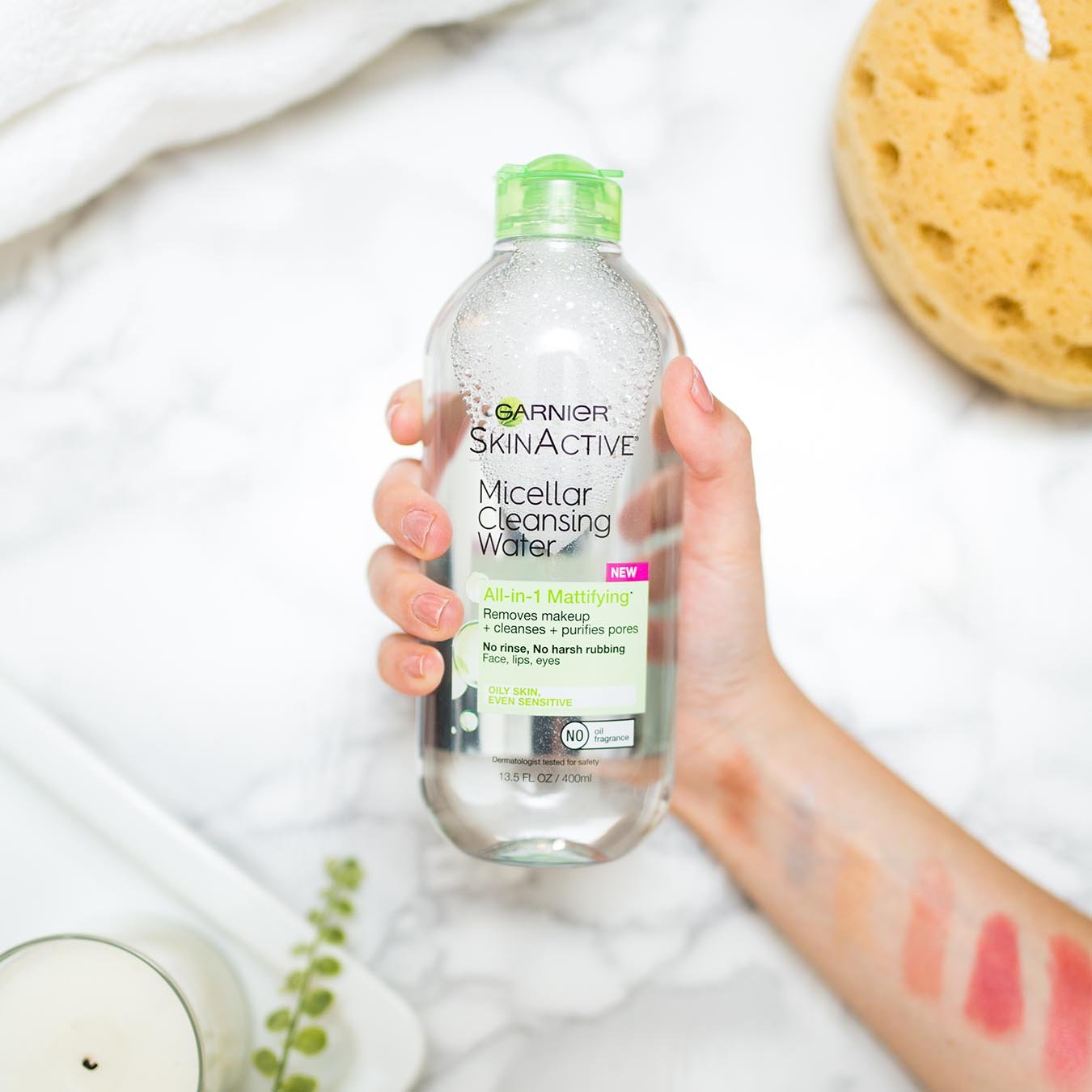 Garnier SkinActive Micellar Cleansing Water All-in-1 Mattifying 13.5 oz with green lid being held by a female hand with 6 shades of makeup swiped across her arm over white marble beside a bath sponge on a white rope, folded white bath towel, and a tray holding a white candle and a eucalyptus branch.